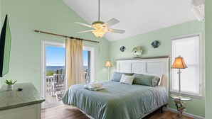 Quarto com cama, ventoinha no teto, janela com persianas, uma cadeira e vista para o oceano.