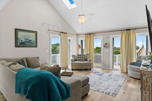 A spacious living area with a sofa, armchair, and ottoman, a ceiling fan, and large windows with curtains. - D by the Sea: 4-bedroom, plus a loft with additional sleep space, oceanfront retreat (Oak Island)