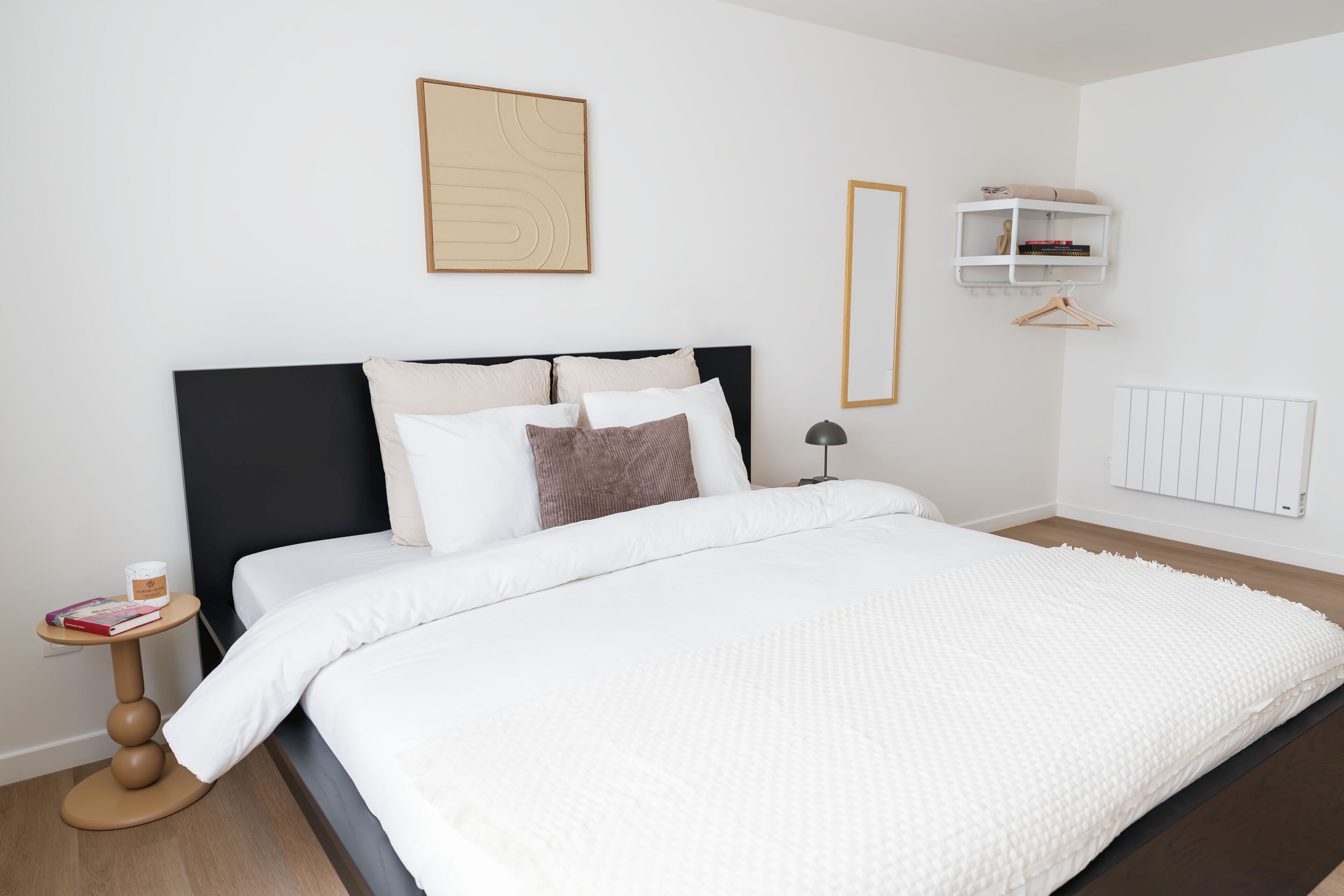 Un lit bien fait, avec une couette blanche et des oreillers, une table de chevet sur laquelle se trouvent un livre et un petit récipient, une étagère murale avec des livres et un cintre, et un radiateur fixé au mur.