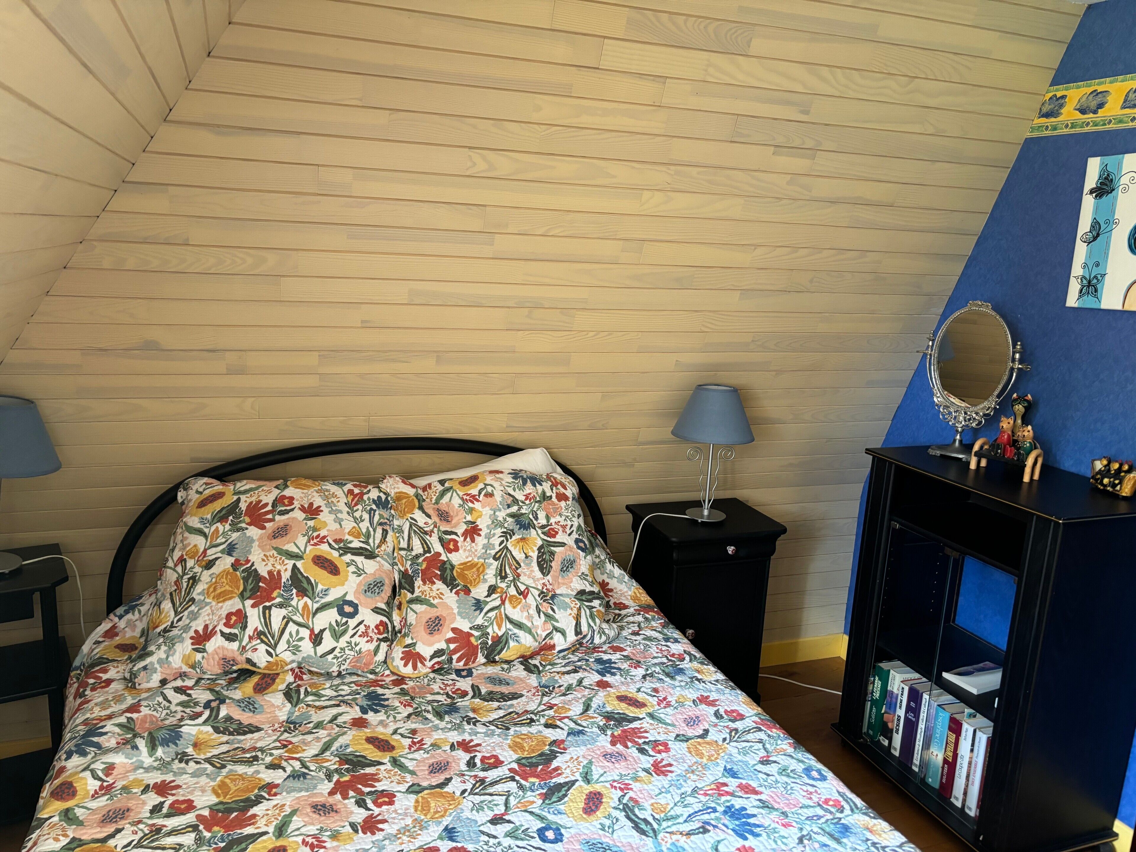 Un dormitorio con techo inclinado, una cama con ropa de cama floral, una mesita de noche con lámpara, un aparador con espejo y una estantería.