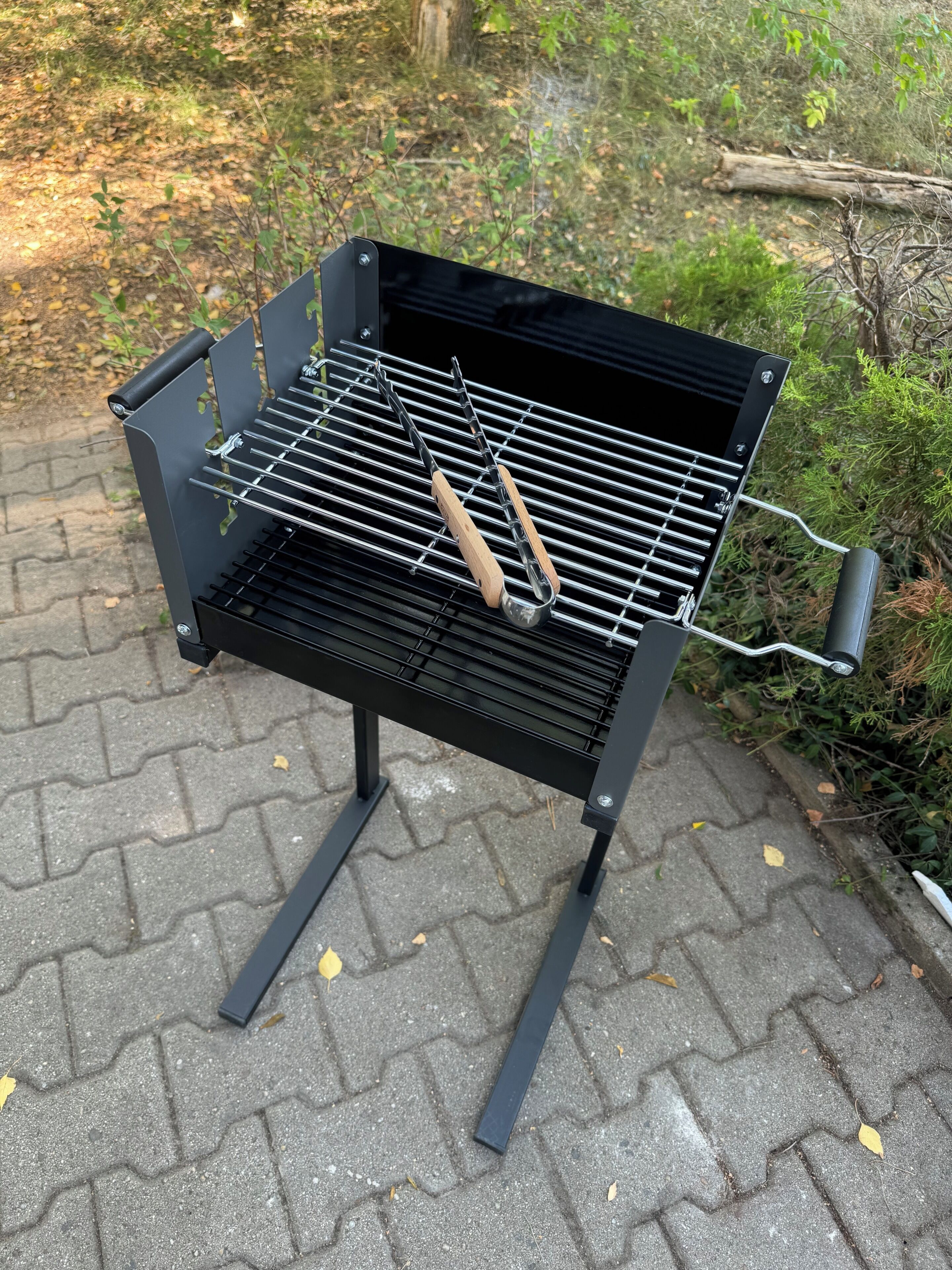 Un barbecue portable doté de grilles en métal et de poignées en bois, placé sur une surface pavée.