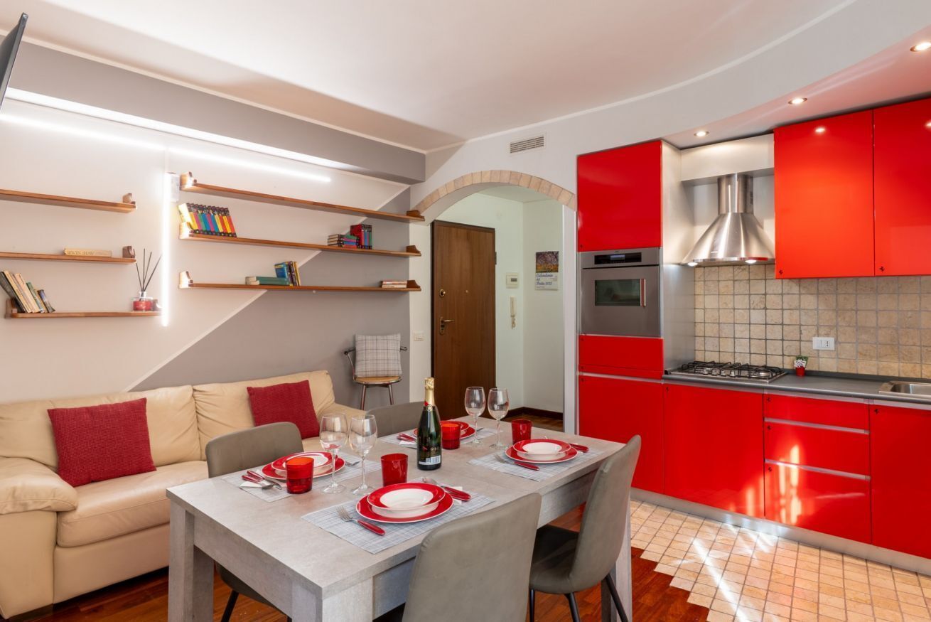Une cuisine moderne avec des meubles rouges, un coin repas avec une table dressée pour quatre personnes, et un salon avec un canapé et des étagères.