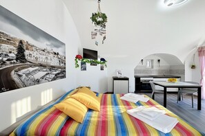 A compact hotel room with a bed, a small desk, a chair, a television, and a kitchen area.