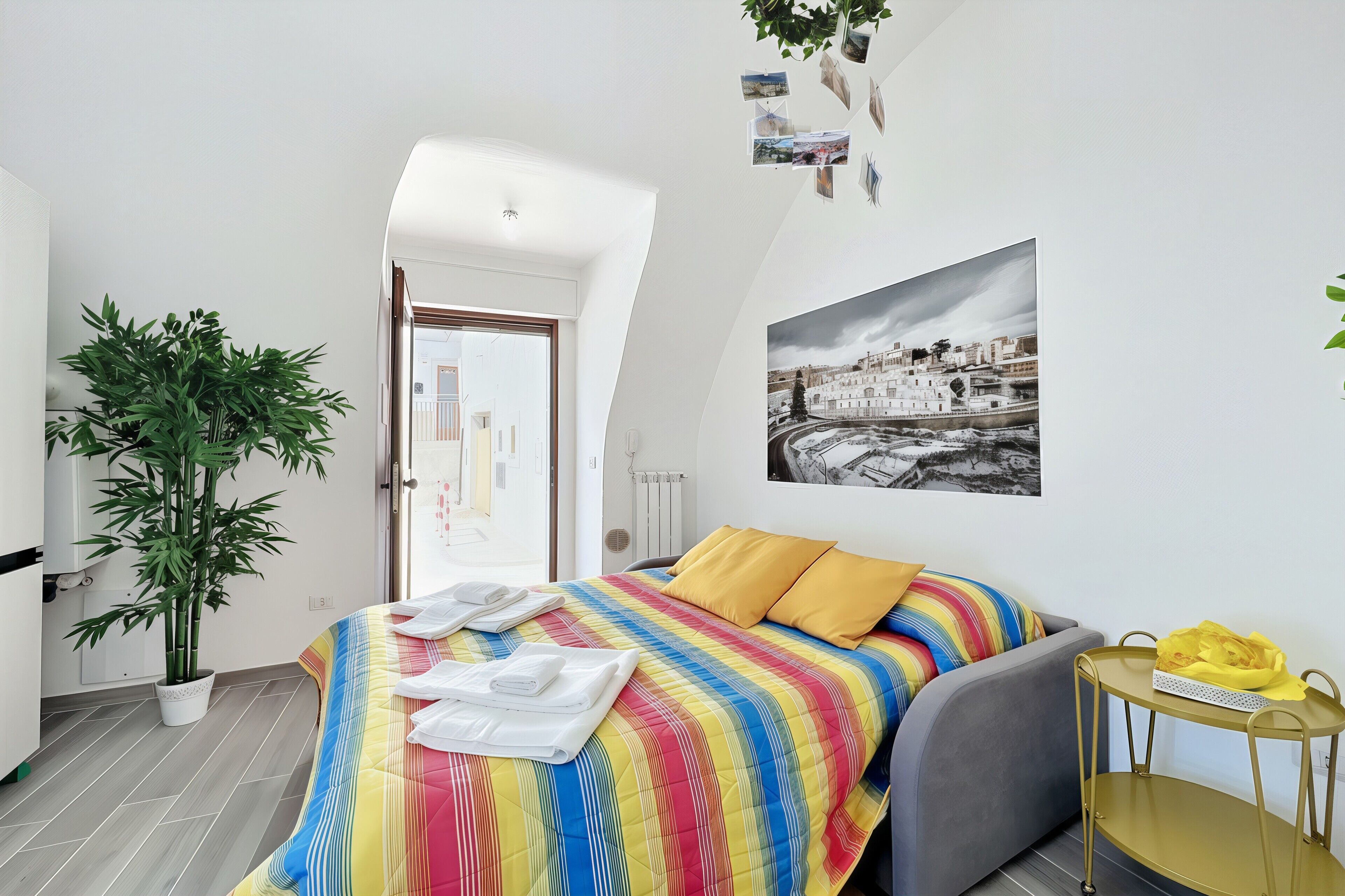 A bedroom with a bed, a potted plant, a wall-mounted photo, and a small table with a yellow object.