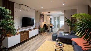 Ein modernes Wohnzimmer mit Flachbildfernseher, einer Couch, einem Couchtisch und einem Essbereich im Hintergrund.