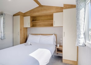 A bedroom with a bed, a nightstand, a wardrobe, and a window with curtains. - Bronze 2 Bedroom Caravan, Sleeps 6 (Walton-on-the-Naze)