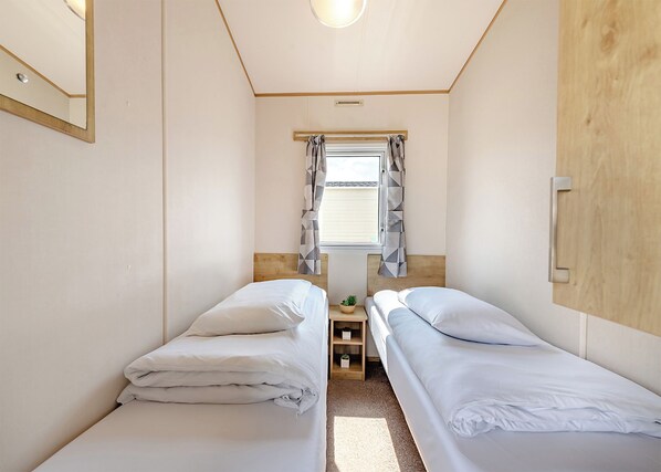 A small, single-room accommodation with two single beds, a window with curtains, a wooden headboard, and a small bedside table with a plant.