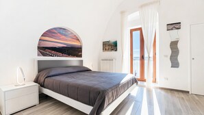 A bedroom with a bed, a nightstand, a wall-mounted mirror, a painting, and a view of the outside.
