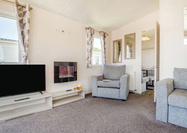 Een moderne woonkamer met een flatscreen-tv, grijze fauteuils, een kleine tafel met een afstandsbediening en een croissant, en een eethoek op de achtergrond.
