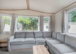 Un sofá seccional gris con una mesa de centro de madera, dos copas de vino y vistas a una zona residencial a través de ventanas grandes con cortinas florales.