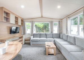 A modern living room with a grey sectional sofa, a TV mounted on a wooden wall, a small wooden table, and large windows with curtains.