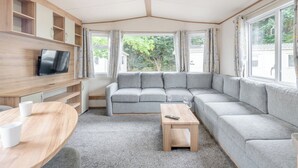 A modern living room with a grey sectional sofa, a TV mounted on a wooden wall, a small wooden table, and large windows with curtains. - Gold Caravan with parking P (Walton-on-the-Naze)