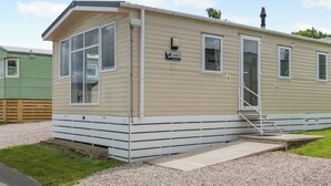 A single-story mobile home with a front porch, a chimney, and a small set of steps leading to the door. - Silver 3 Bedroom Caravan, Sleeps 8 P (Walton-on-the-Naze)