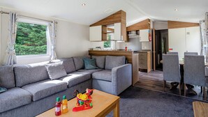 A modern interior with a grey sofa, wooden accents, a dining area, and a view of greenery outside. - Silver Premium 2 Bedroom Caravan, Sleeps 6 P (Walton-on-the-Naze)