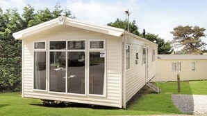 A small, single-story mobile home with a pitched roof, large windows, and a visible door. - Silver Premium 2 Bedroom Caravan, Sleeps 6 P (Walton-on-the-Naze)