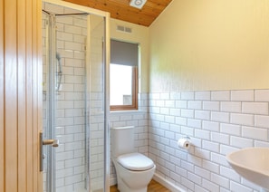 Une salle de bain avec des murs recouverts de carreaux blancs, des toilettes, une douche en verre et une fenêtre.