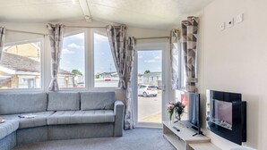 A living room with a grey sofa, a flat-screen TV, a window with curtains, and a view of the outside. - Gold Accessible with parking P (Walton-on-the-Naze)