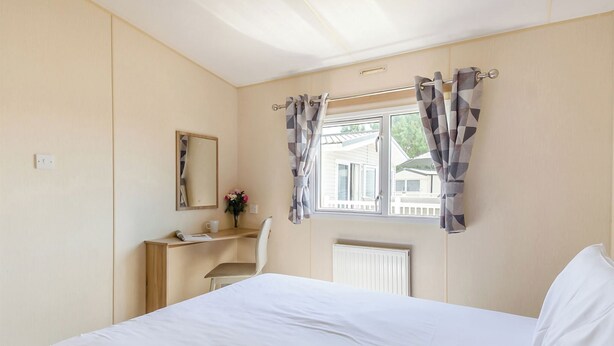 Een slaapkamer met een bed, bureau, stoel, raam met gordijnen, radiator en spiegel.