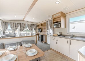 A compact kitchen with a microwave, toaster, and sink, a dining area with a table set for two, and a TV mounted on the wall. - Gold 3 Bedroom Caravan, Sleeps 8 (Ryde, Nr Bembridge)