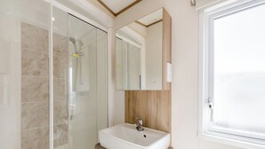 Ein Badezimmer mit Duschkabine aus Glas, einem weißen Waschbecken mit Holzplatte, einem Spiegel über dem Waschbecken und einem Fenster zur Rechten.