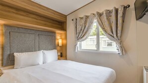 Ein Hotelzimmer mit Bett, Kopfteil, Fenster mit Vorhängen, einem an der Wand befestigten Fernseher und einem Nachttisch.