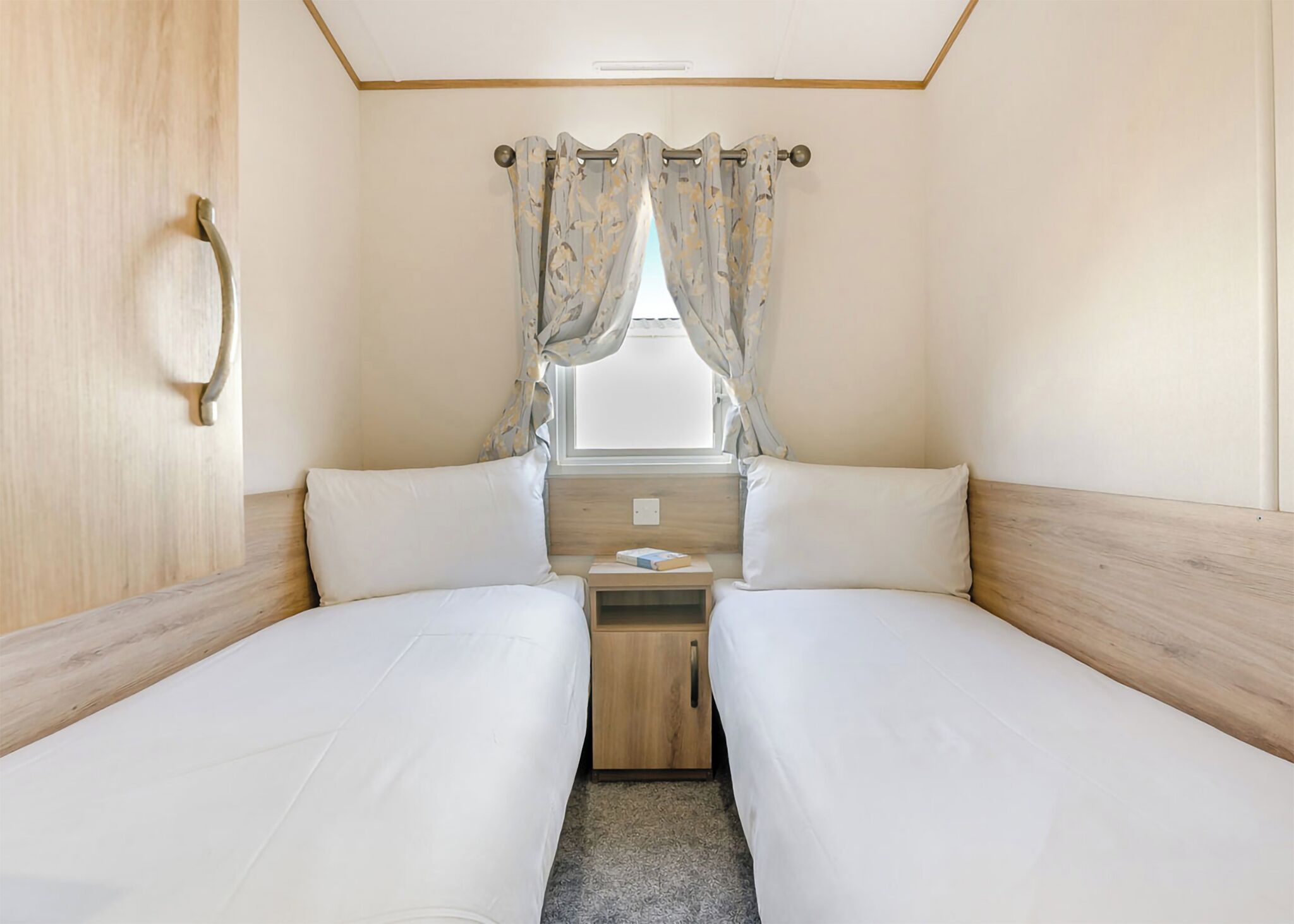 Ein Doppelzimmer mit zwei Betten, einem hölzernen Kopfteil, einem kleinen Nachttisch und einem Fenster mit Vorhängen.