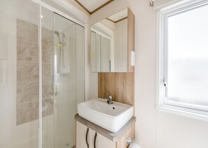 A bathroom with a glass shower enclosure, a white sink with a wooden countertop, a mirror above the sink, and a window to the right. - Gold Caravan with parking & decking (Ryde, Nr Bembridge)