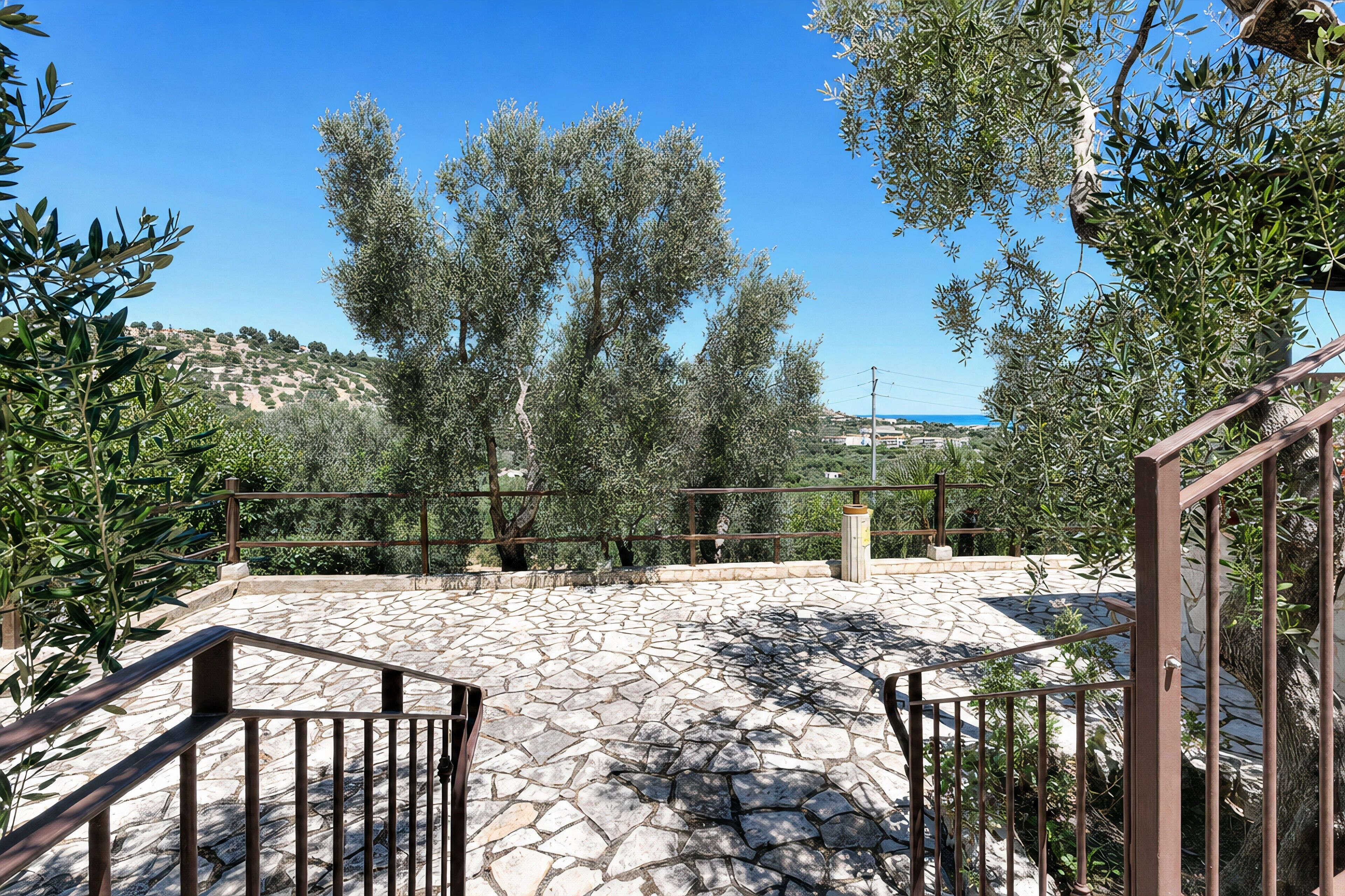 A stone patio with metal railings, olive trees, and a view of a hilly landscape.