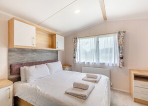 A bedroom with a bed, bedside tables, a wardrobe, a window with curtains, and a radiator. - Gold Premium Caravan with ensuite & decking (Ryde, Nr Bembridge)