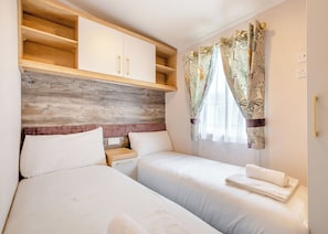 A hotel room with two beds, wooden headboard, a small wooden shelf, and a window with curtains. - Gold Premium Caravan parking, ensuite, deck SV 6 P (Ryde, Nr Bembridge)