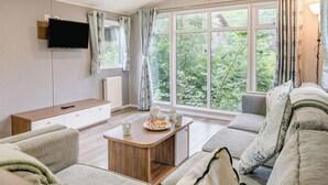 A modern living room with a flat-screen TV, a wooden coffee table, a sofa, and a large window with curtains. - Gold Premium Caravan, parking, ensuite, deck & SV (Ryde, Nr Bembridge)