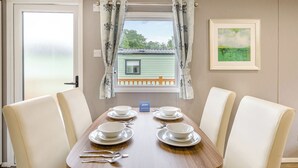 A dining area with a table set for four, a window with curtains, and a framed picture on the wall. - Gold Premium Caravan, parking, ensuite, deck & SV (Ryde, Nr Bembridge)