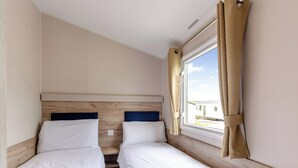 A room with two beds, a window with curtains, and a view of buildings outside. - Silver 3 Bedroom Caravan, Sleeps 8 (Ryde, Nr Bembridge)