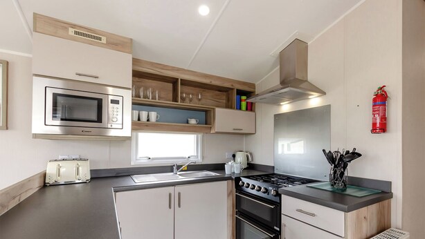 A compact kitchen with a microwave, toaster, sink, stove, and wooden shelves. - Silver 3 Bedroom Caravan, Sleeps 8 (Ryde, Nr Bembridge)