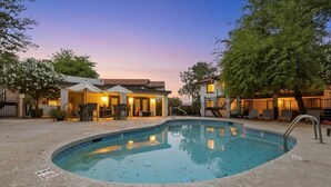A swimming pool with a tiled deck, a covered patio area with chairs, and a building with a red roof. - Great for Mid/Long Stays - Pool - King Bed (Mesa)