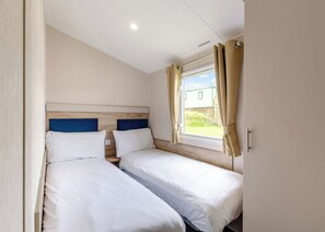 A room with two beds, a window with curtains, a wooden headboard, and a light switch on the wall. - Silver Caravan with decking P (Ryde, Nr Bembridge)