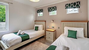 A bedroom with two beds, a nightstand, a lamp, and a window with patterned curtains. - Deluxe Lodge with parking, ensuite & decking 4 (Ryde, Nr Bembridge)