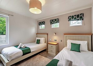 A bedroom with two beds, a nightstand, a lamp, and a window with patterned curtains. - Deluxe Lodge with parking, ensuite & decking 4 (Ryde, Nr Bembridge)
