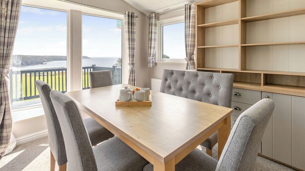 A dining area with a wooden table, grey upholstered chairs, and a view of the sea through large windows. - Deluxe Lodge, parking, ensuite, decking & sea view (Ryde, Nr Bembridge)