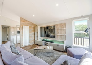 A modern living room with a sofa, coffee table, and a kitchen area with a large window and a balcony. - Prestige Lodge, parking, ensuite, decking & SV 4 (Ryde, Nr Bembridge)