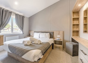 A modern bedroom with a wooden bed frame, a bedside table with a lamp, a small desk, and a built-in shelving unit. - Prestige Lodge with parking, ensuite & decking 4 (Ryde, Nr Bembridge)