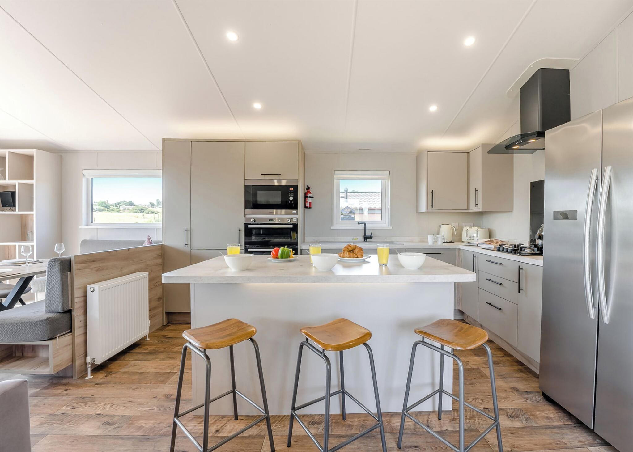 Una cocina moderna con una isla central, taburetes de bar, electrodomésticos de acero inoxidable y vistas al exterior.