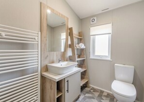 A bathroom with a white sink, mirror, and toilet, wooden shelves, and a radiator. - Prestige Lodge with parking, ensuite & decking 4 (Ryde, Nr Bembridge)
