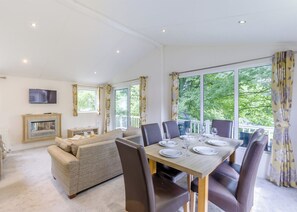 A dining area with a table set for four, a sofa, a fireplace, and large windows with curtains. - Prestige Lodge, parking, ensuite, decking & SV 6 (Ryde, Nr Bembridge)