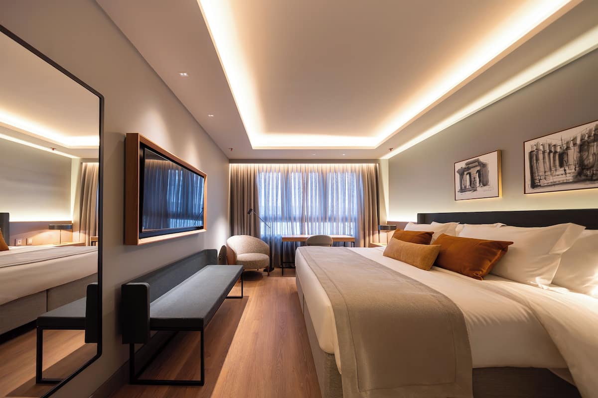 A modern hotel room with a large bed, a desk, a chair, and a mirror.