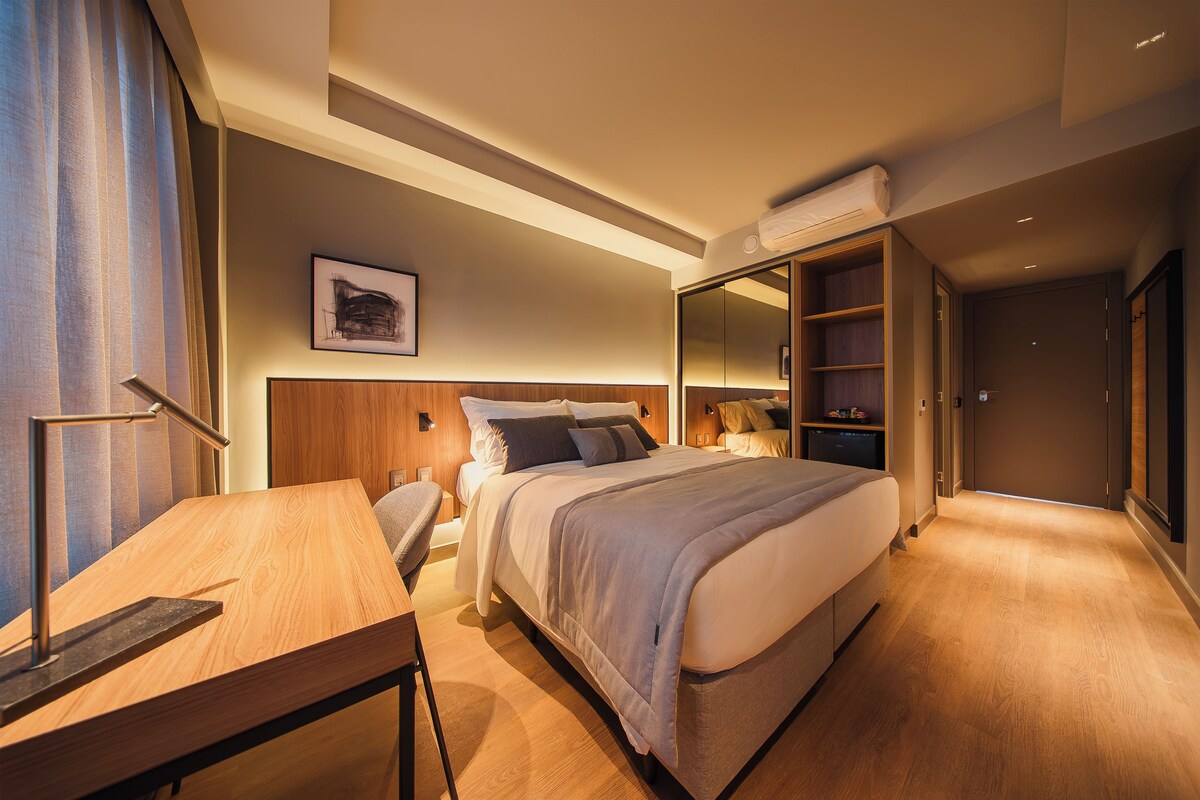 A modern hotel room with a bed, a desk, a chair, a wardrobe, and a mirror.