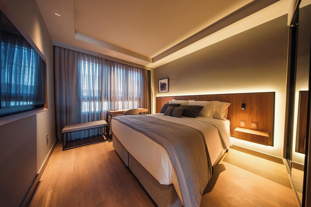 A modern hotel room with a large bed, bedside tables, a bench, and a view of the outdoors.