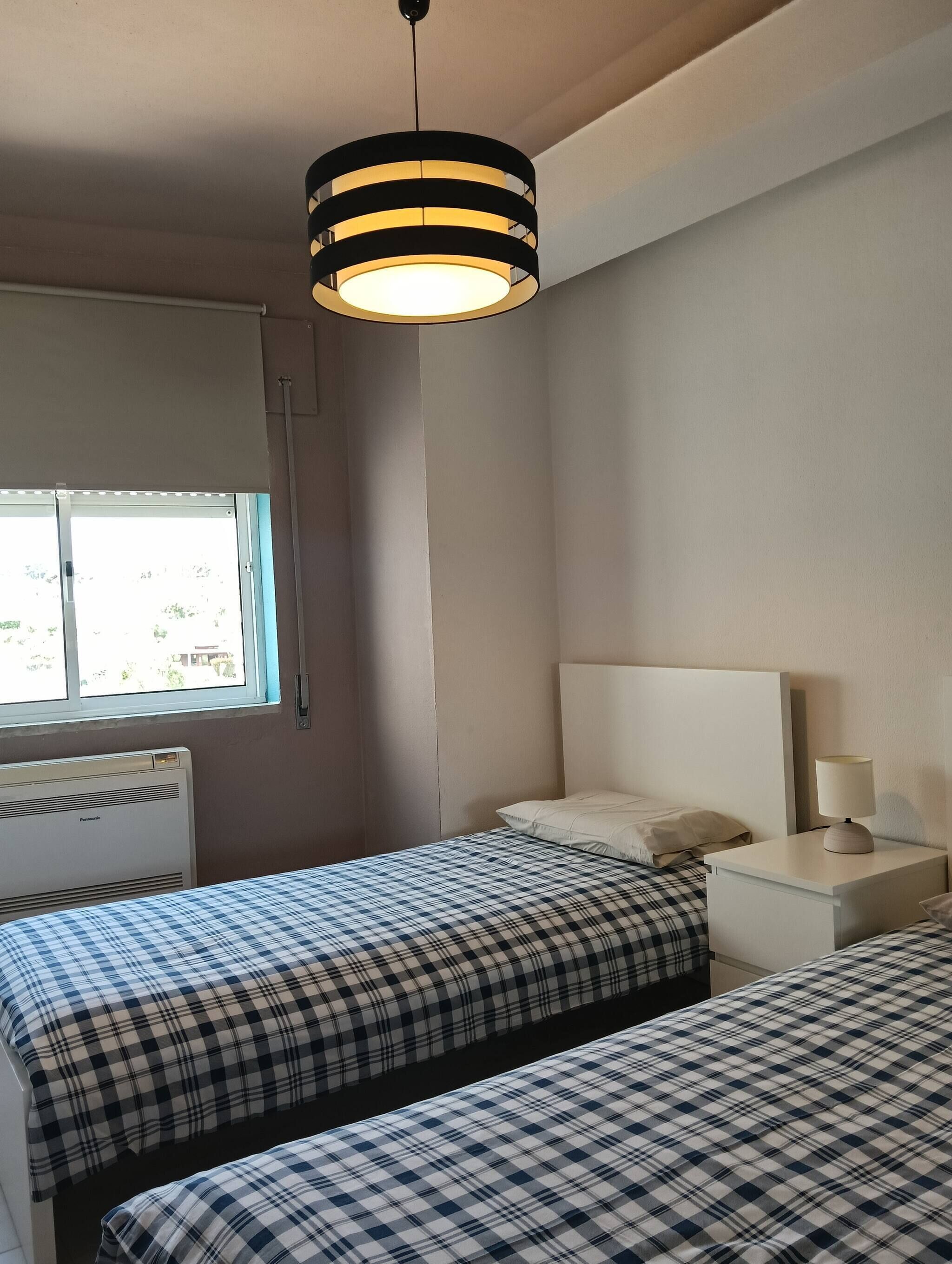 A room with two beds, a window, a radiator, and a hanging light fixture.