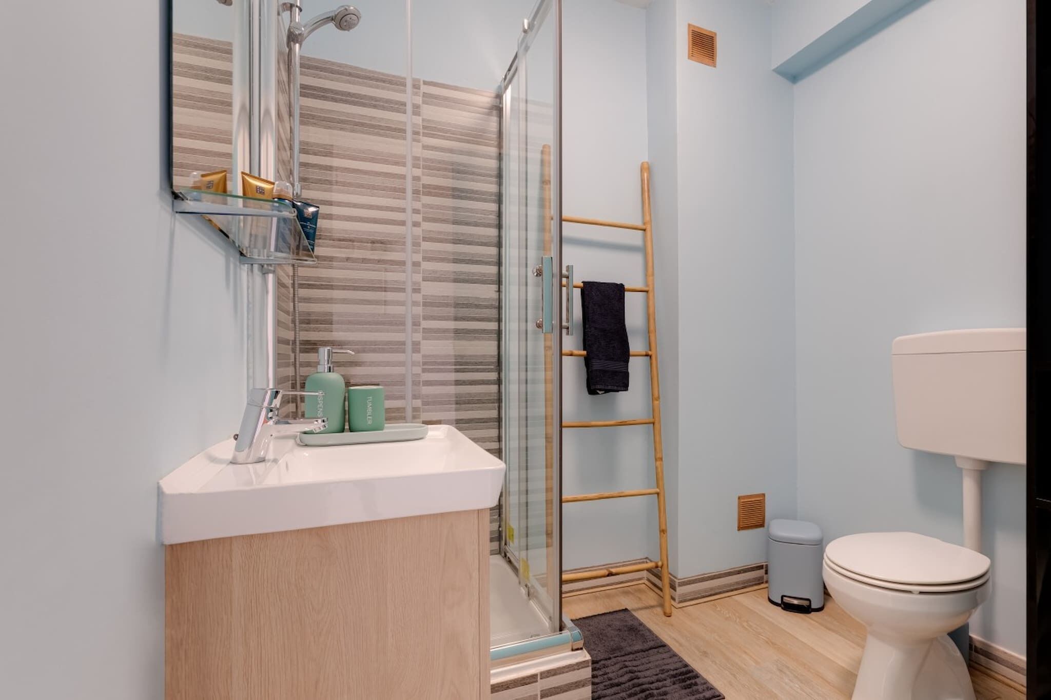 Une salle de bain moderne avec une douche à l’italienne en verre, une échelle en bois, des toilettes blanches et un lavabo blanc avec un miroir au-dessus.