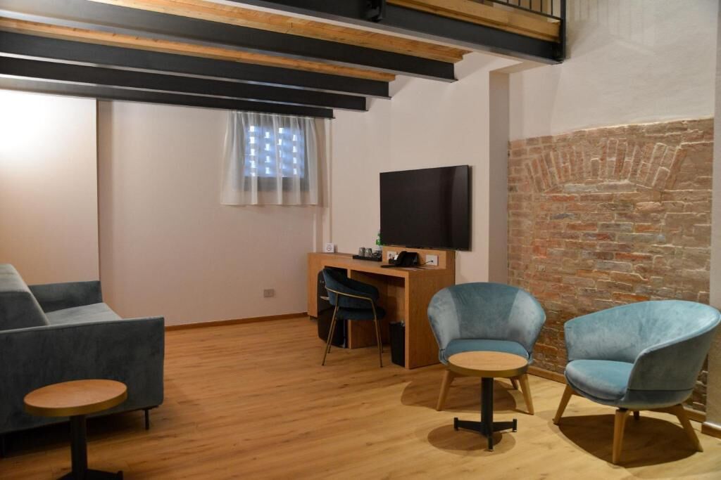 Una sala de estar moderna con piso de madera, un sofá, dos sillones, una pequeña mesa redonda, un escritorio con una computadora y un televisor de pantalla plana, además de una pared de ladrillo.